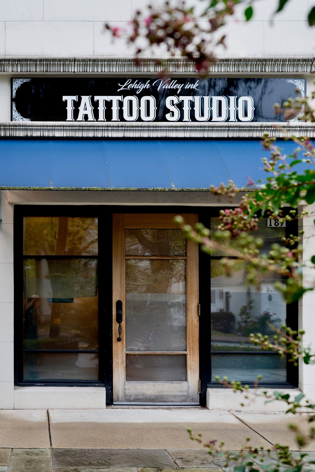 Exterior view of a tattoo studio with a sign above the entrance, partially obscured by branches.